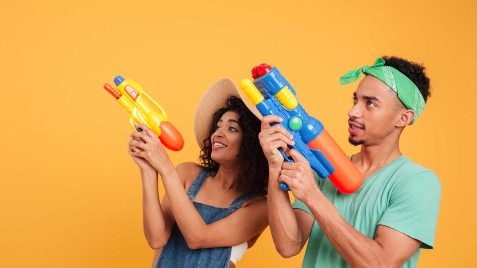 Portrait of a cheerful young african couple dressed in summer clothes playing with water guns isolated over yellow background
