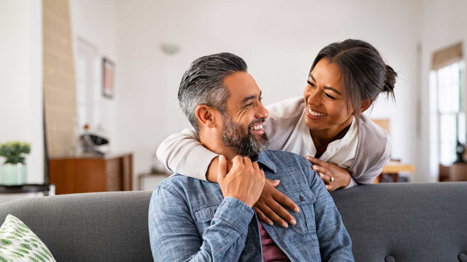 Smiling ethnic woman hugging her husband on the couch from behind in the living room. Middle eastern man having fun with his beautiful young wife on the couch. Mid adult indian man with latin woman.