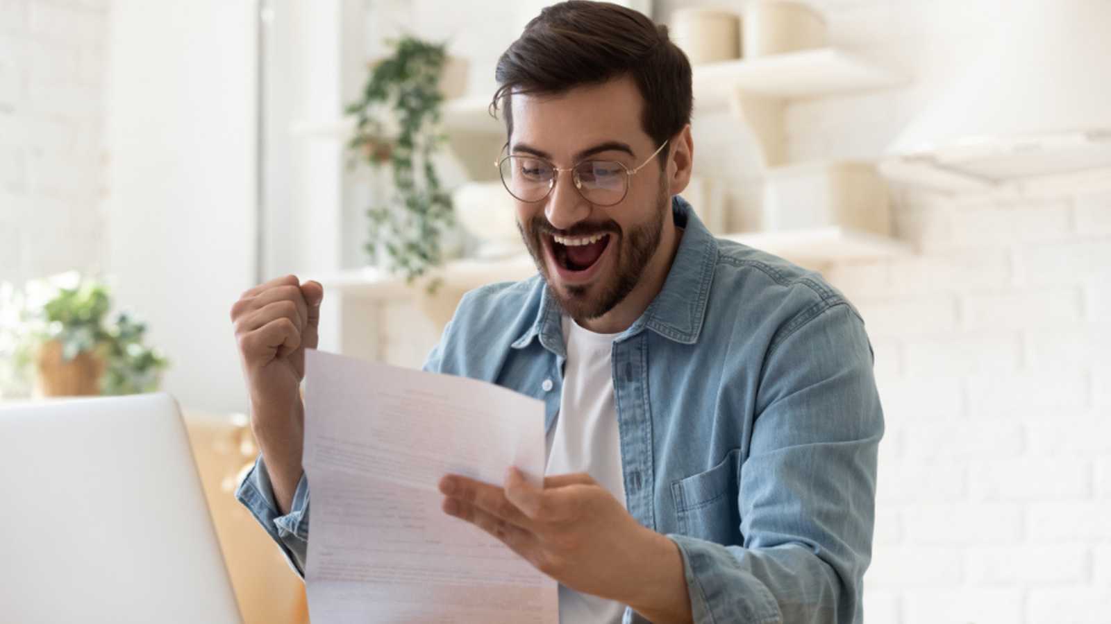 Excited euphoric happy young man holding reading paper postal mail letter amazed overjoyed by good news, got new job celebrate taxes refund receive salary payment loan approval sit at home table