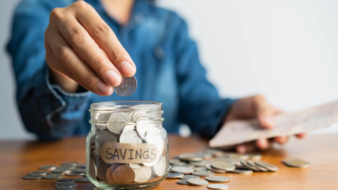 asian woman hand is putting a coin in a glass bottle and a pile of coins on a brown wooden table,Investment business, retirement, finance and saving money