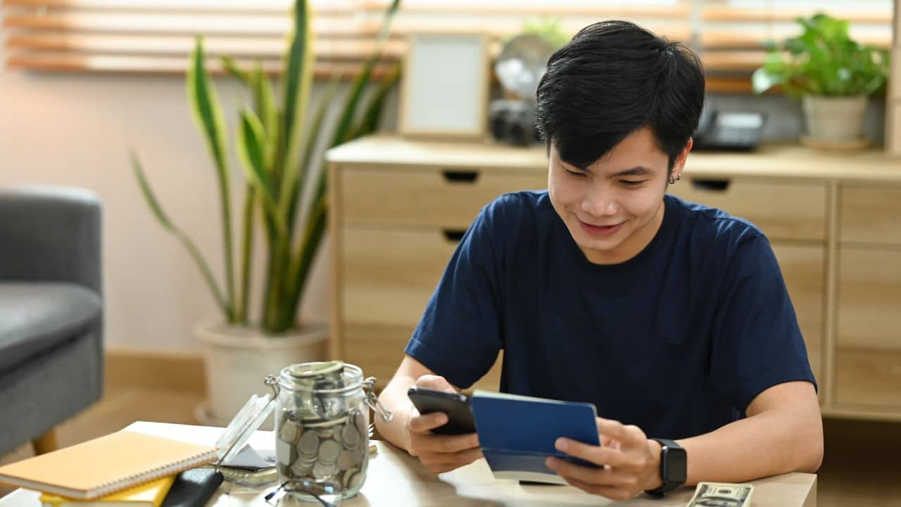 Satisfied asian man using smart phone for making online payments, managing expenses finances in living room