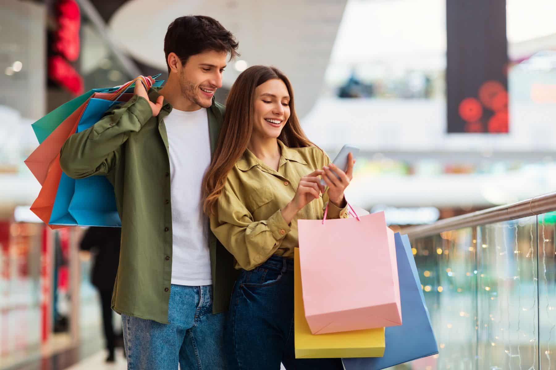 Buyers Couple Shopping Using Cellphone Holding Colorful Shopper Bags Standing In Mall. 