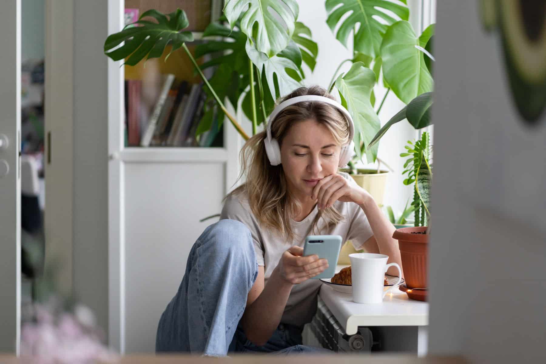 Woman listening to music, wear wireless white headphones, using mobile smart phone, chatting in social networks, sitting next to the window, houseplants on windowsill.