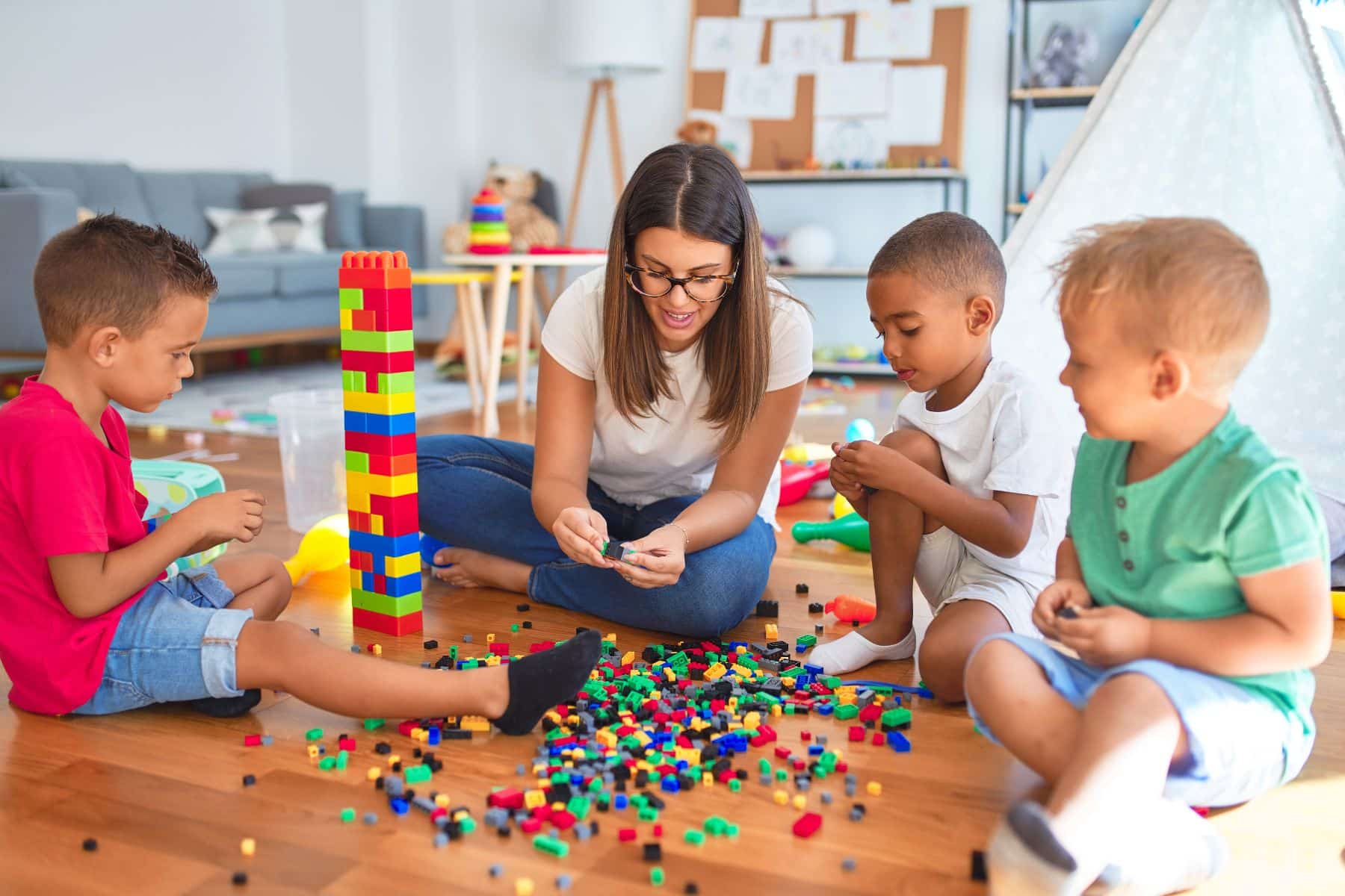 Young beautiful teacher and toddlers playing with building blocks around lots of toys at kindergarten
